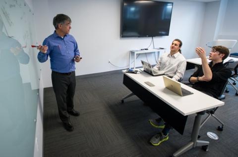 在UNH创业中心，两个人拿着笔记本电脑坐在一张桌子旁，一个人站在一块白板旁