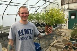 温室里的男学生拿着一株植物. 他的t恤上写着UNH研究生院.